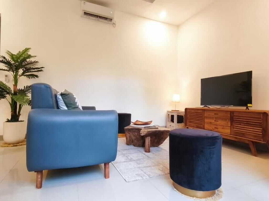 A living room with a blue sofa, a round ottoman, a wooden coffee table, a TV, and a potted plant.