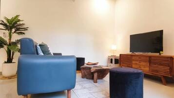 A living room with a blue sofa, a round ottoman, a wooden coffee table, a TV, and a potted plant.