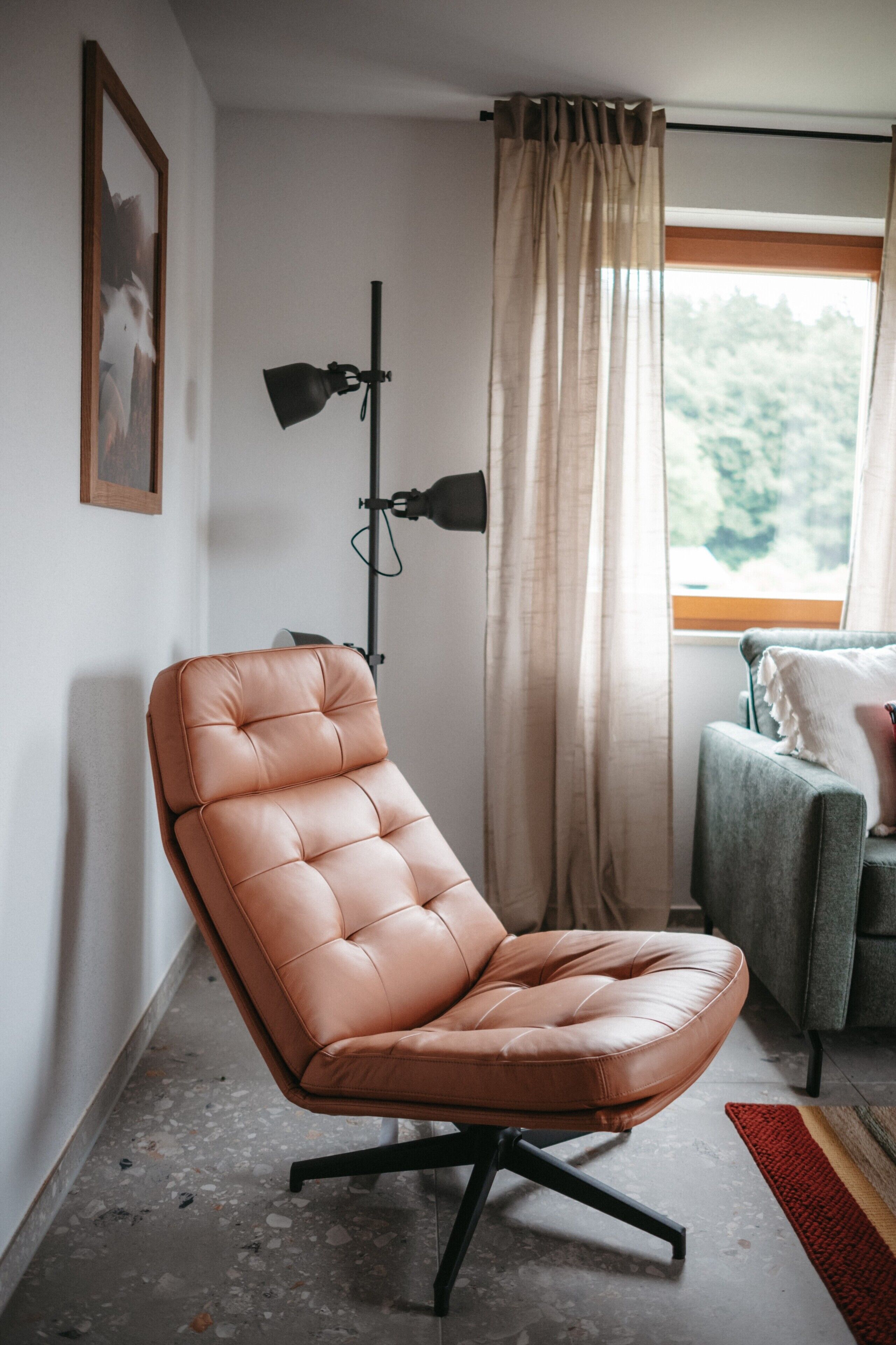 Een kamer met een leren fauteuil, een vloerlamp, een bank, een raam met gordijnen en een ingelijst schilderij aan de muur.