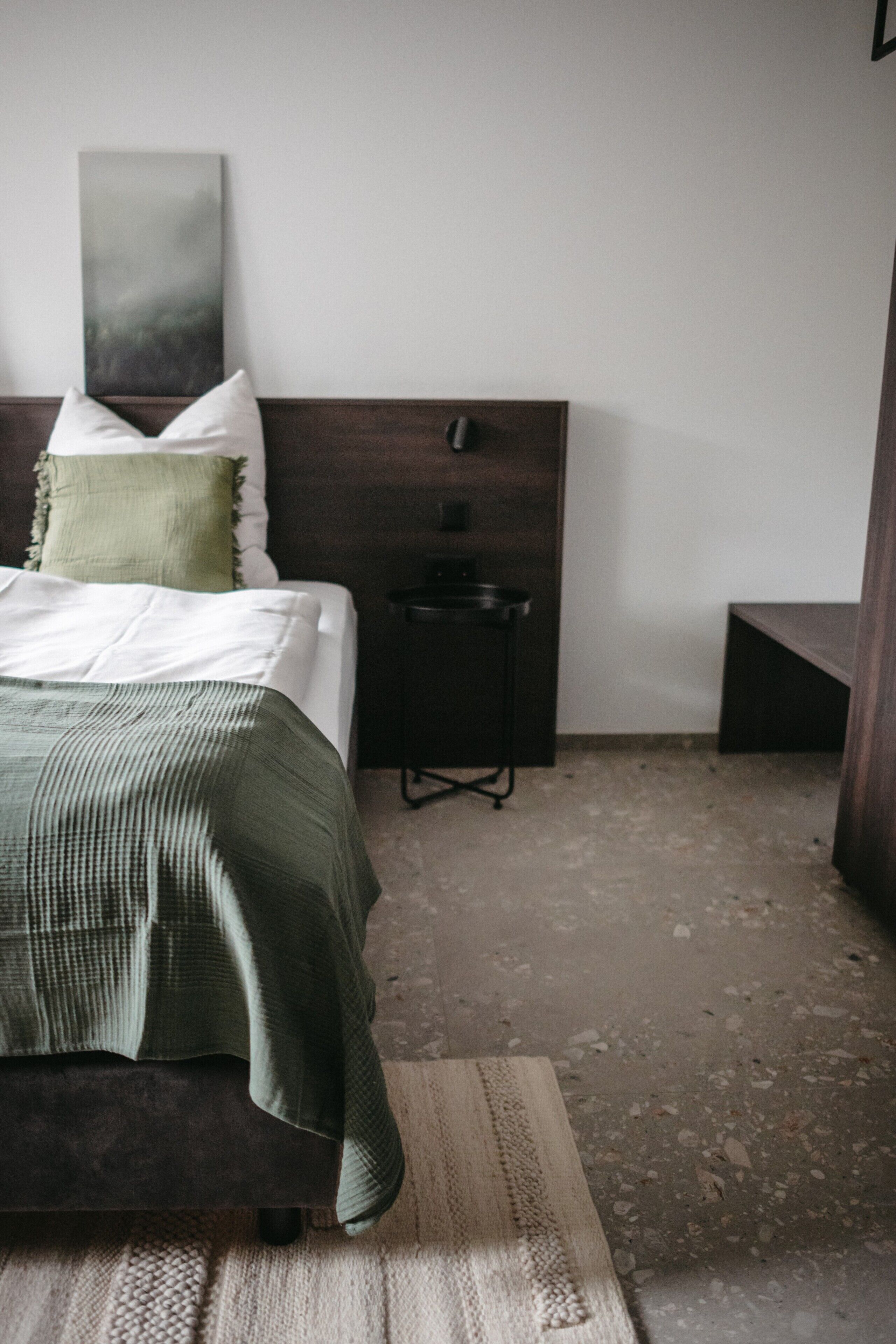 A bedroom with a bed, a bedside table, a nightstand, a wall-mounted artwork, and a concrete floor.