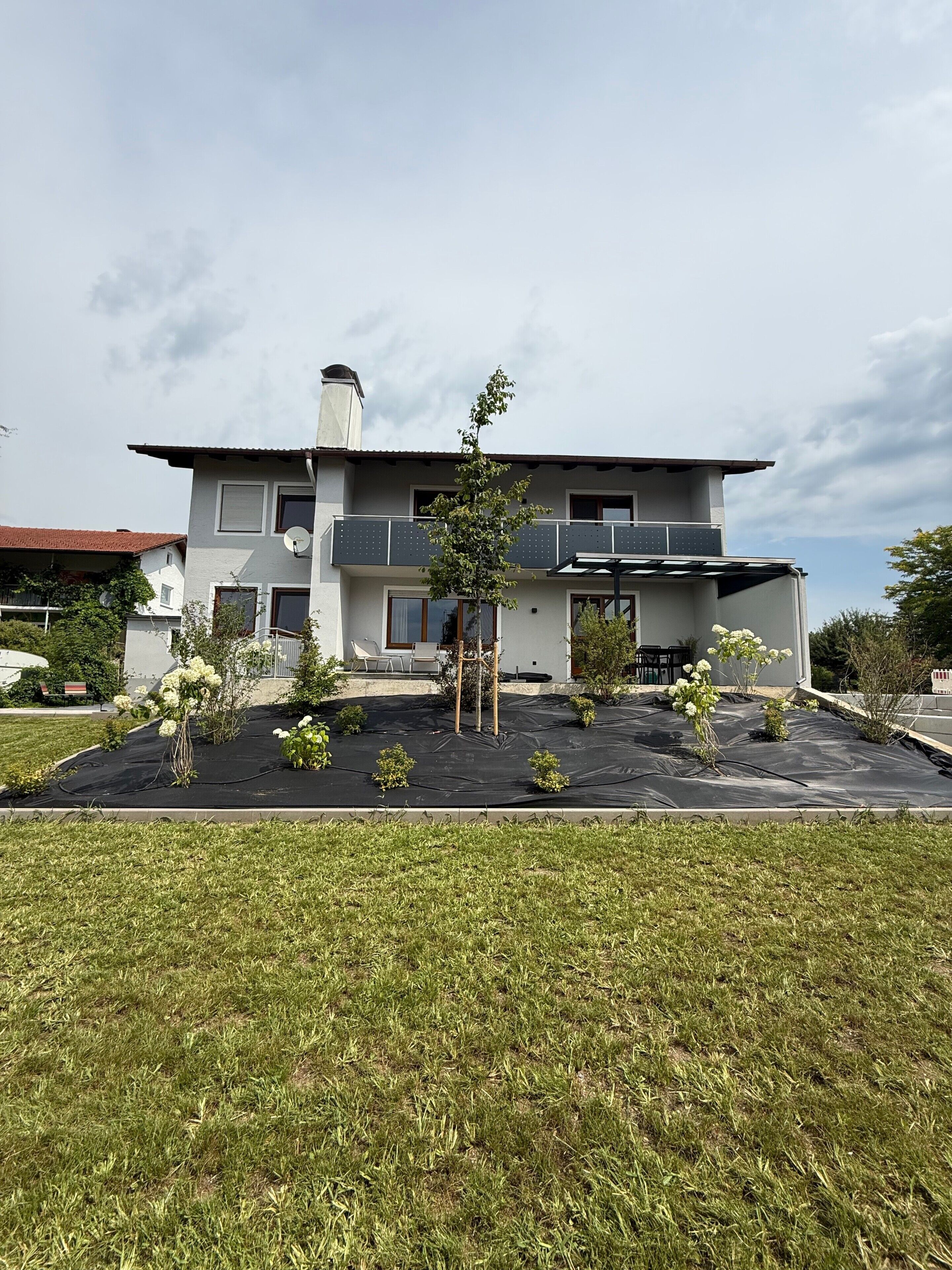 Ein modernes, zweistöckiges Haus mit flachem Dach, einem Balkon und einem Gartenbereich mit verschiedenen Pflanzen.