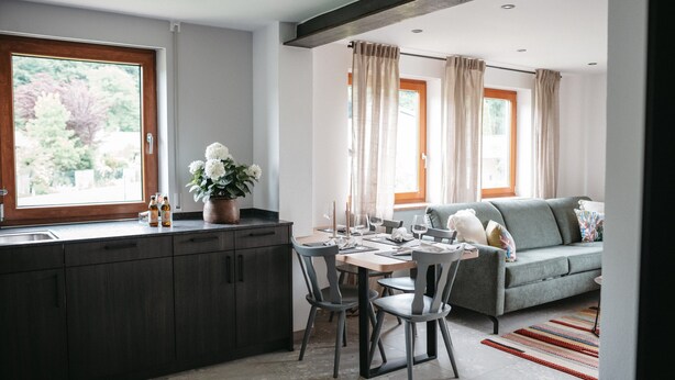 A modern kitchen with dark cabinets, a dining area with a table set for four, and a grey sofa with a view of the outdoors.