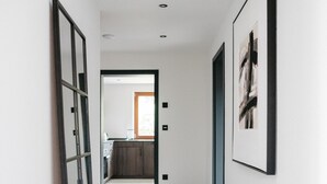 A hallway with a kitchen in the background, framed artwork on the walls, and a tiled floor.