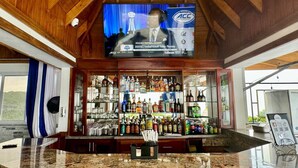 Zona de bar con una televisión mostrando el inicio del fútbol ACC, estantes llenos de diversas botellas y una barra con un tablero de menú.