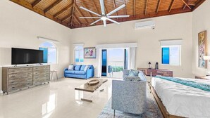 Una habitación amplia con techo alto, una cama grande, un sofá azul, televisión y vistas al exterior.
