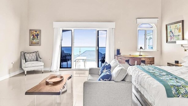 Una habitación amplia con ventilador de techo, cama con cubrecama floral, sofá con almohadas, bandeja de madera y ventana grande con vistas.