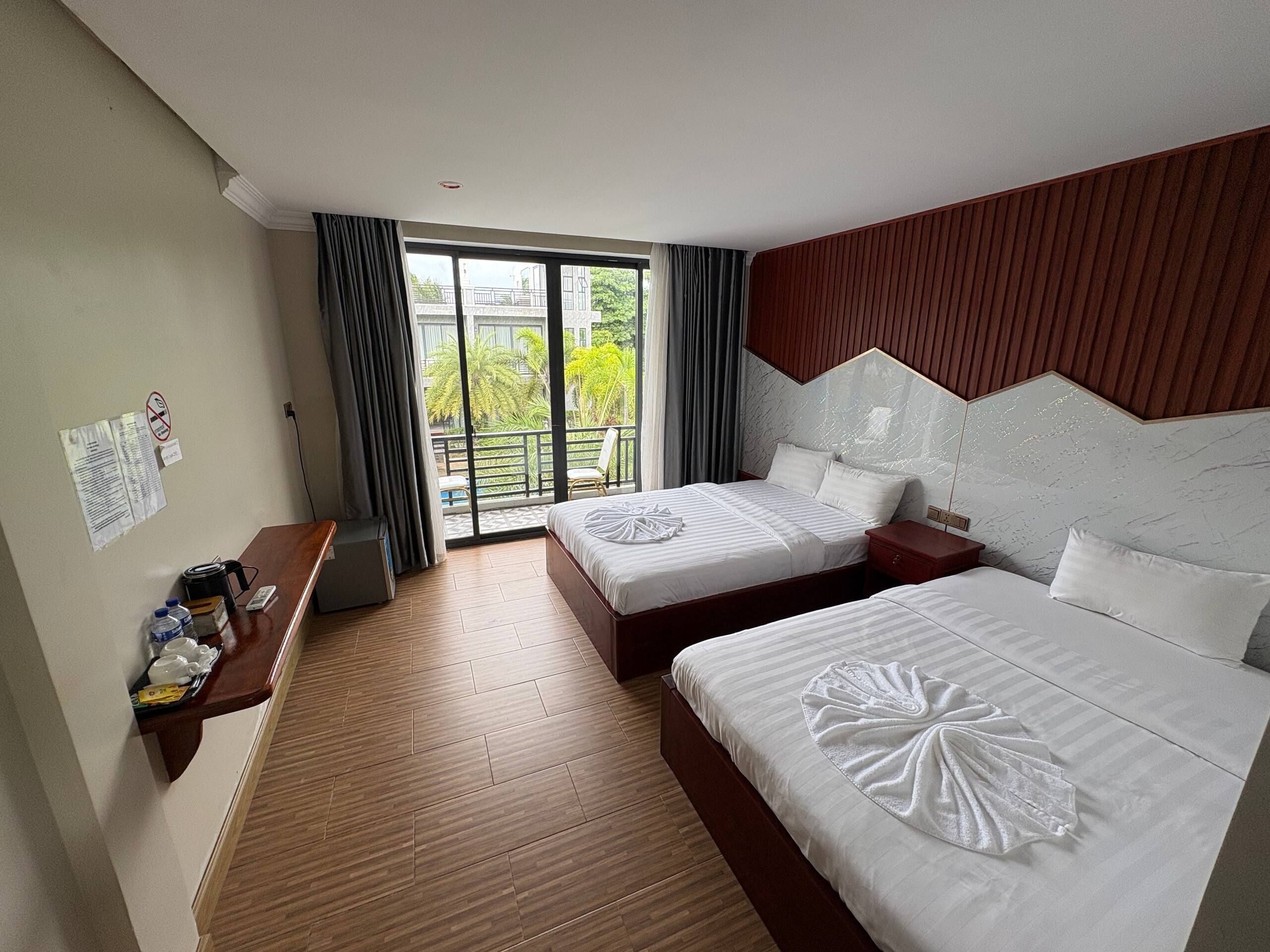 A hotel room with two beds, a wooden floor, a balcony with a view, and a desk with a chair.