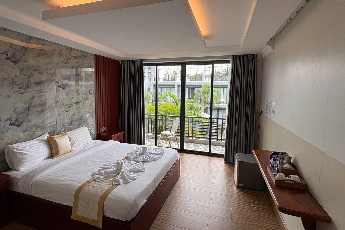 A hotel room with a large bed, a desk with a kettle, and a view of a building and greenery outside.