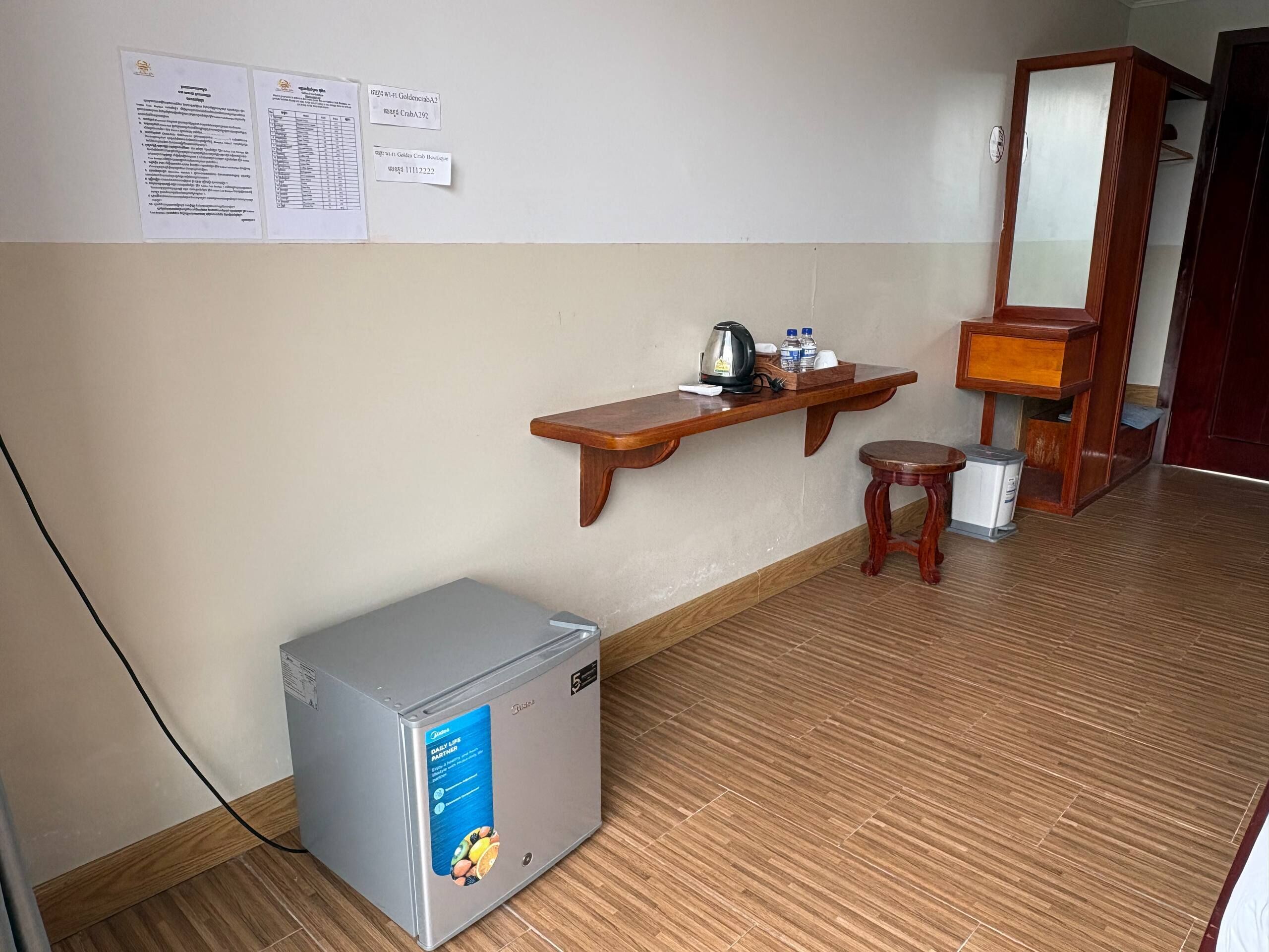 A compact room with a wooden floor, a small refrigerator, a wooden shelf with a kettle and water bottles, a wooden stool, and a mirror.