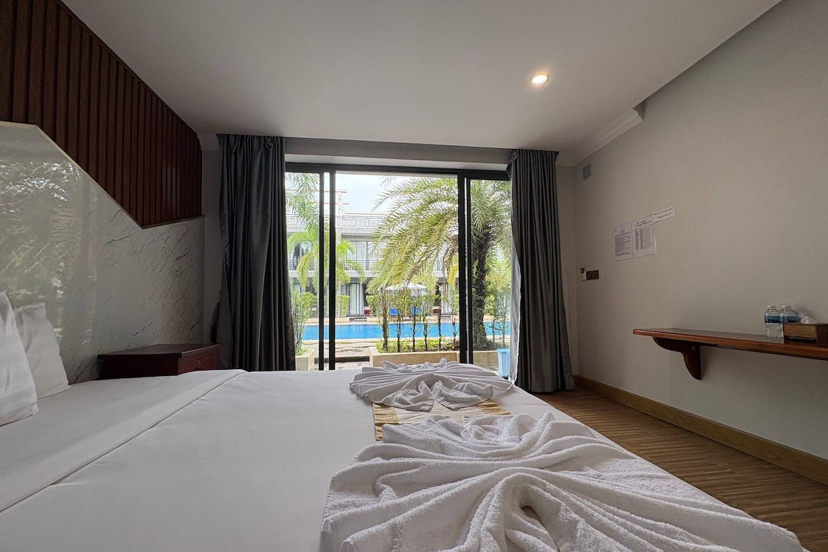 A hotel room with a bed, a wooden nightstand, a window with curtains, a view of a swimming pool, and a wooden shelf with items.