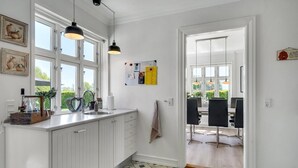 Una cocina moderna con armarios blancos, un lavamanos y luces colgantes. Se ve un área de comedor con una mesa y sillas a través de una puerta abierta.