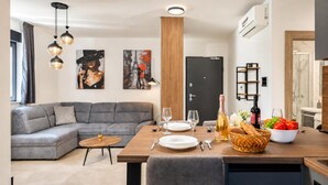 A modern living room with a grey sectional sofa, a wooden dining table set with plates, glasses, and a bottle of wine, and a fruit bowl on the table. - Zadar Family Residence (Zadar)
