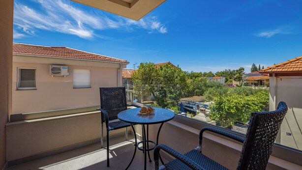 A balcony with two wicker chairs, a small round table, and a view of neighboring buildings and trees. - Zadar Family Residence (Zadar)