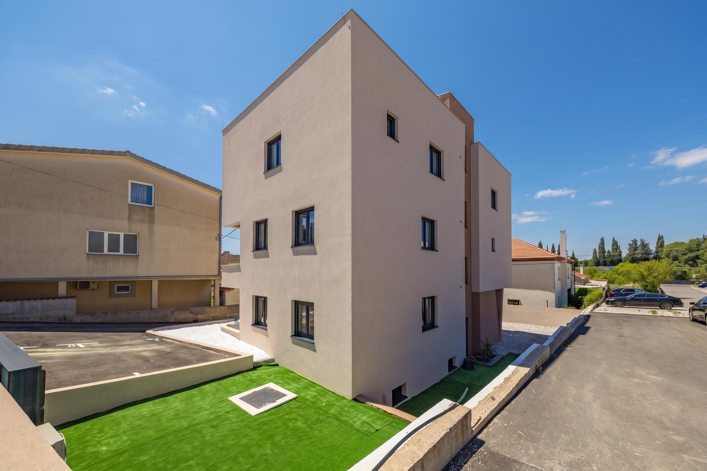 A modern multi-story residential building with a flat roof, multiple windows, and a small green lawn area.