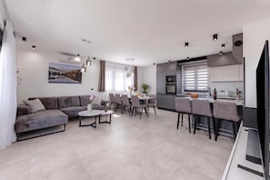 A modern living room with a grey sofa, a dining area with a round table, and a kitchen with a built-in oven and microwave. - Zadar Family Residence (Zadar)