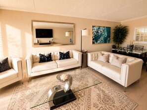 A living room with two white sofas, a glass coffee table, a large abstract painting, a floor lamp, and a dining area with a table and chairs.