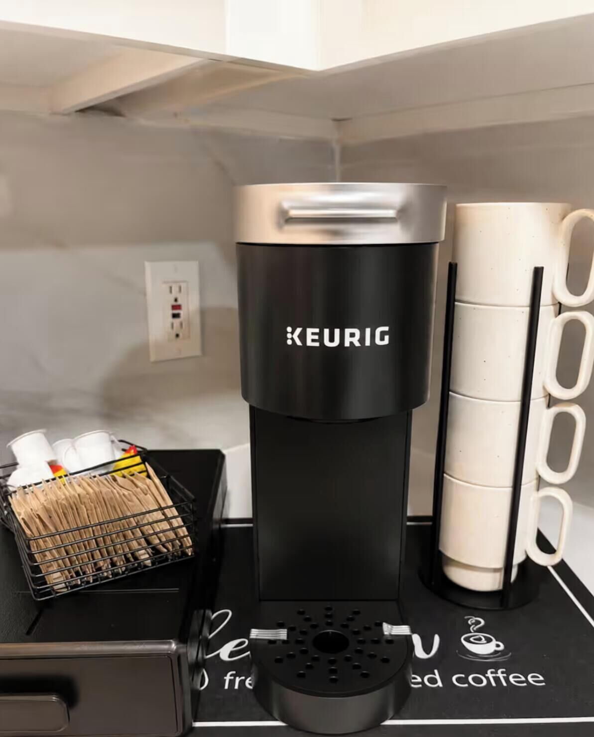 A black Keurig coffee maker on a countertop with a coffee mug holder and a tray with utensils.