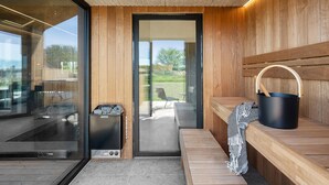 Un sauna avec un banc en bois, un seau noir et une porte en verre donnant sur un espace extérieur.