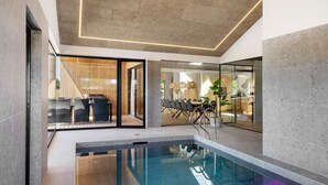 Un espace piscine intérieur moderne avec des murs en verre transparent, un coin repas avec des meubles en bois et une cuisine visible à travers des portes en verre.