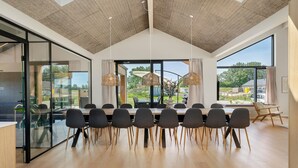 Un espace repas moderne avec une longue table, des chaises et de grandes fenêtres donnant sur l’extérieur.