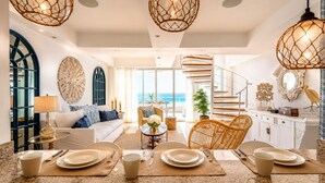 Un amplio salón con vistas al océano, una zona de comedor con platos y cubiertos, y una escalera que conduce a un nivel superior.