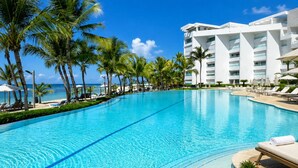 Una piscina azul clara con sillas de descanso, palmeras y un edificio blanco con balcones.