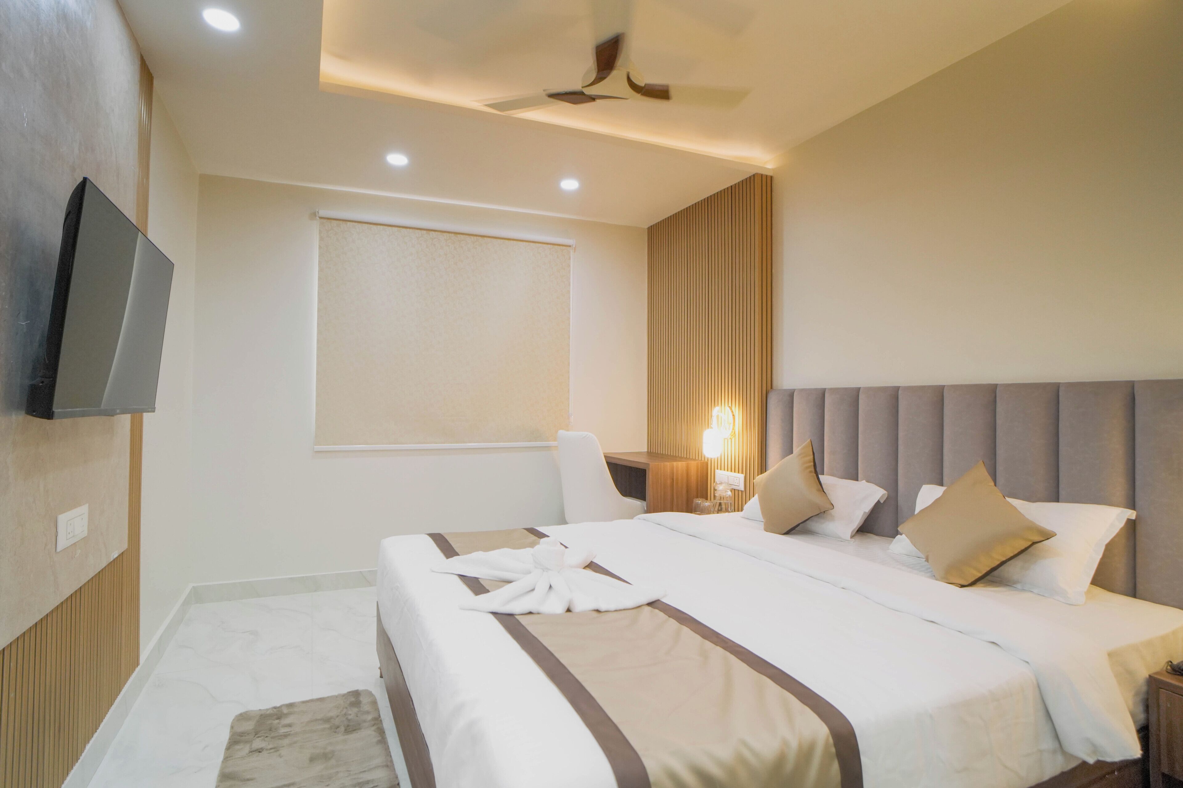 A hotel room with a bed, a flat-screen TV mounted on the wall, a ceiling fan, and a window with a blind.