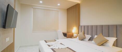A hotel room with a bed, a flat-screen TV mounted on the wall, a ceiling fan, and a window with a blind.