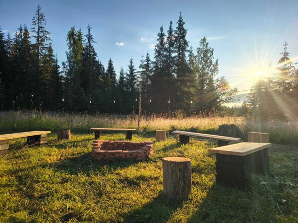 Un espace extérieur aménagé avec des bancs et un foyer, entouré de pelouse et d’arbres.