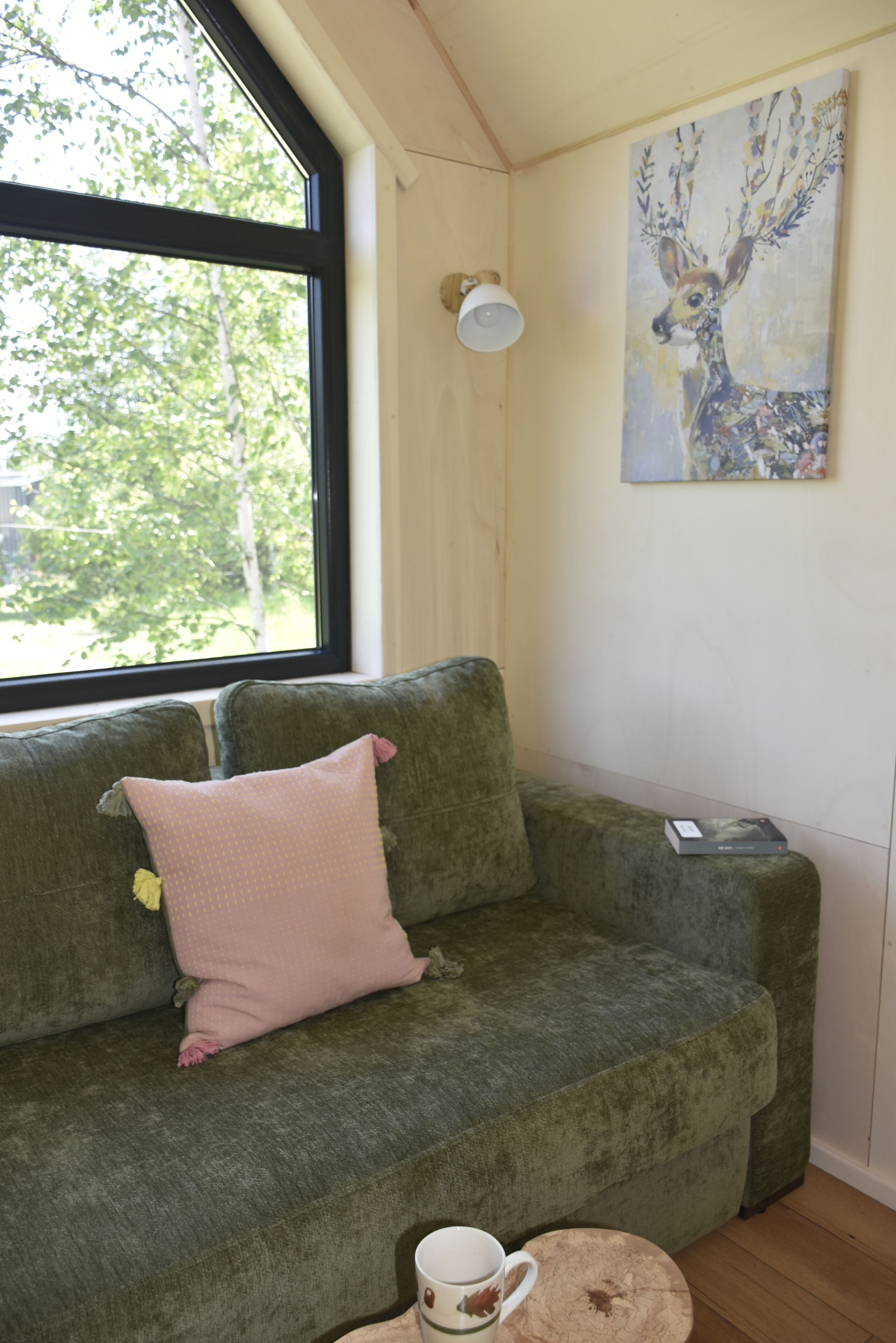 Un sofá verde con un cojín rosa, una taza blanca y una mesita con un libro. En la pared, un cuadro de un ciervo.