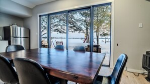 Un espace repas comprenant une grande table, des chaises, un réfrigérateur et une vue sur une étendue d’eau grâce à de grandes fenêtres.