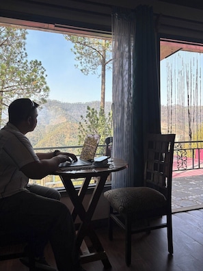 A person seated at a table with a laptop, near a window with a view of trees and hills. - Rustic Pines (Solan)