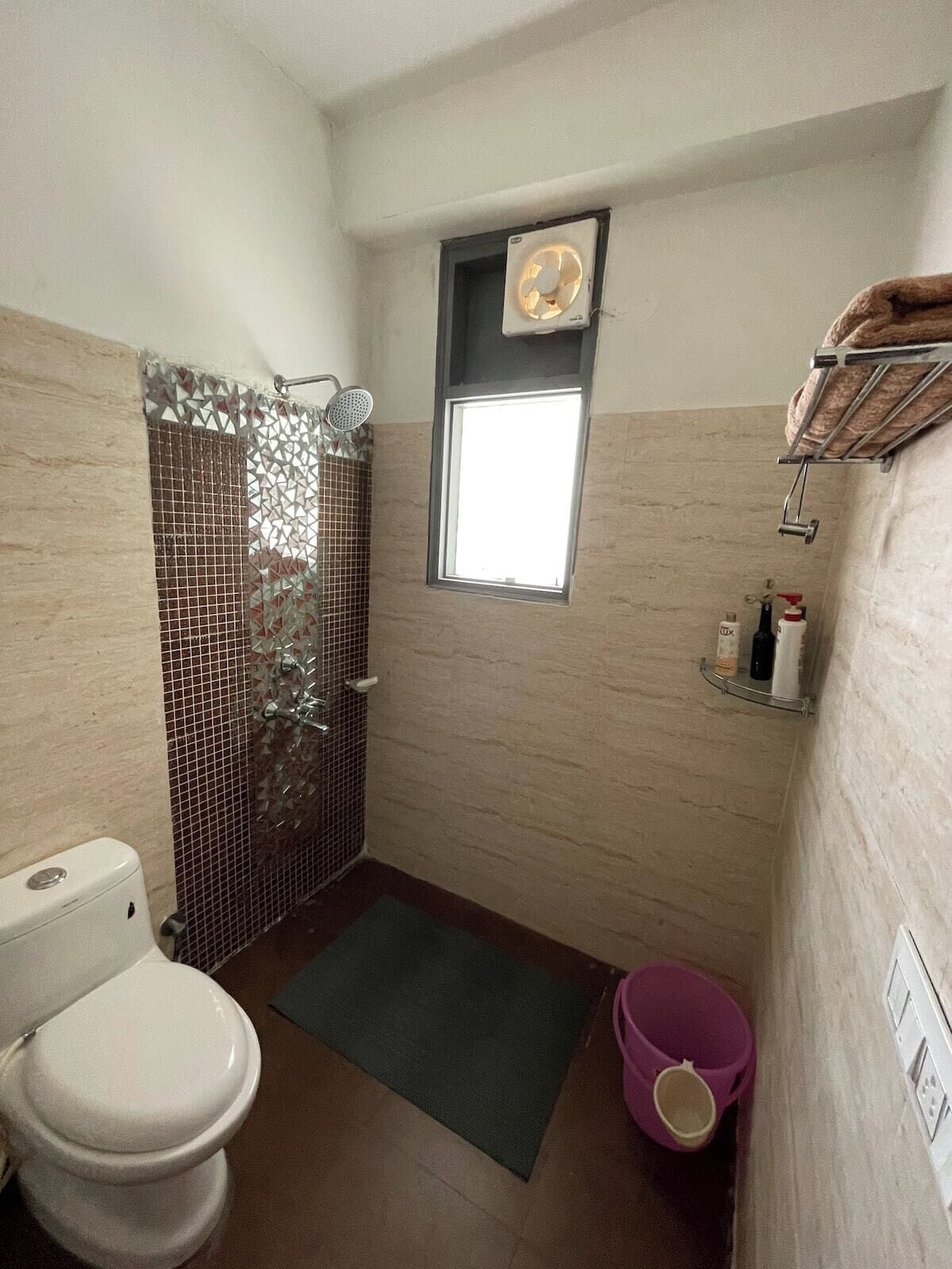 Um banheiro pequeno com vaso sanitário, chuveiro com porta de vidro, janela e prateleira com produtos de higiene pessoal.