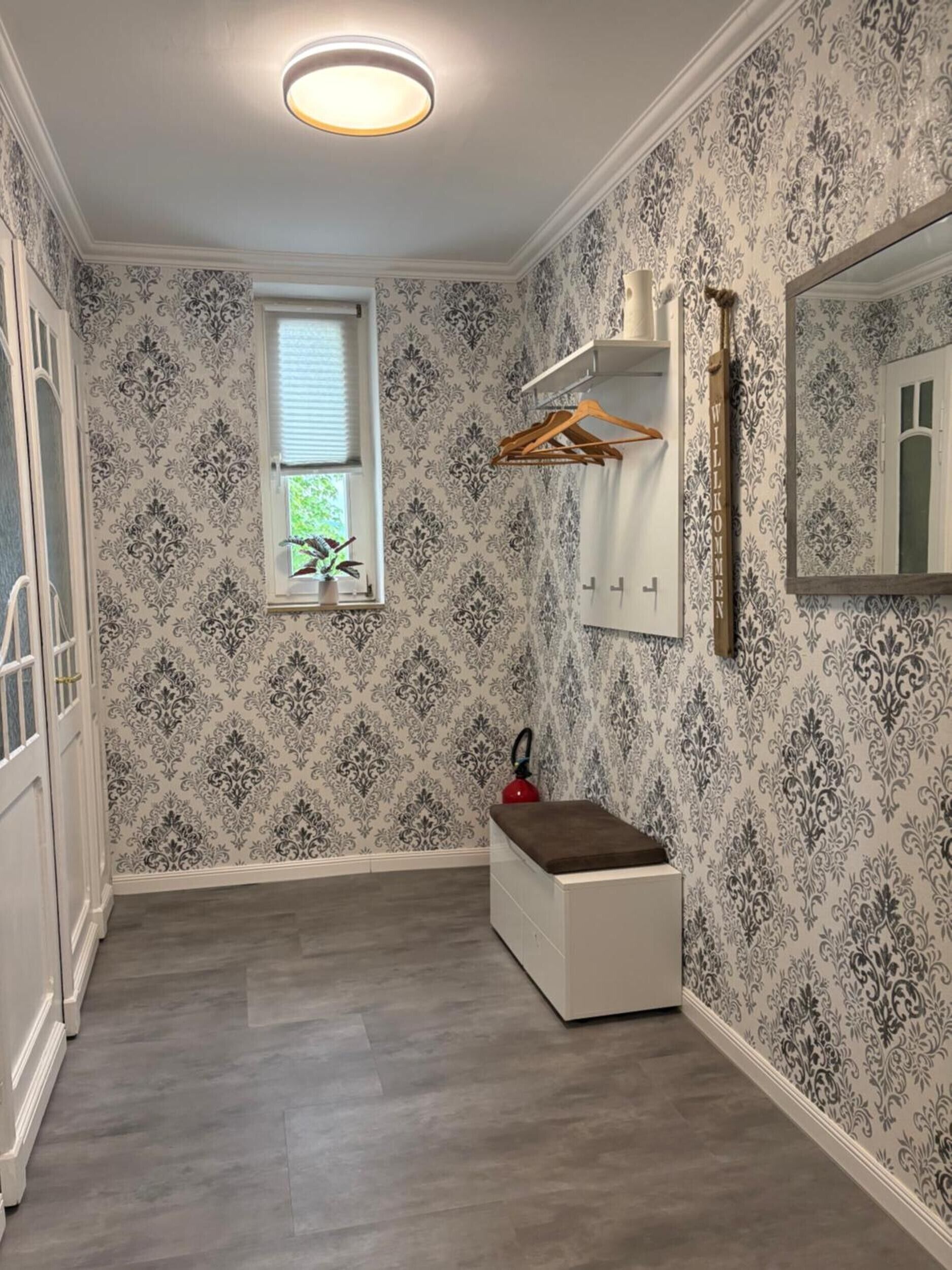 A hallway with patterned wallpaper, a white bench, a fire extinguisher, a window with blinds, and a mirror on the wall.
