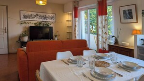 En matplats med ett bord för fyra personer, en soffa, en tv och ett fönster med gardiner.
