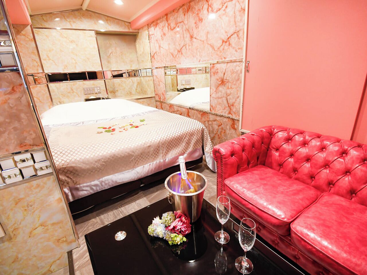 A hotel room with a bed, a red leather sofa, a black coffee table with a bottle of champagne, and a vase of flowers.