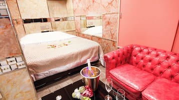 A hotel room with a bed, a red leather sofa, a black coffee table with a bottle of champagne, and a vase of flowers.