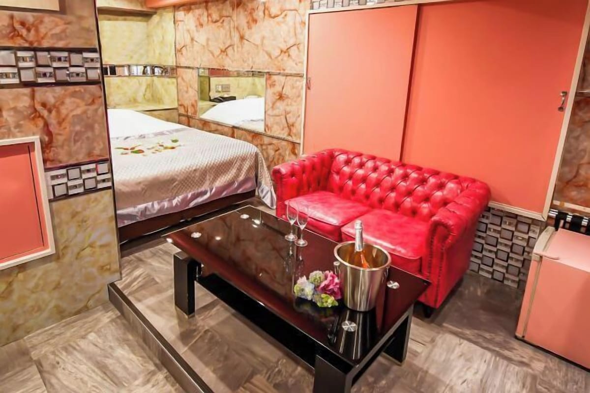 A hotel room with a bed, a red leather sofa, a small table with a bottle, and a pink refrigerator.