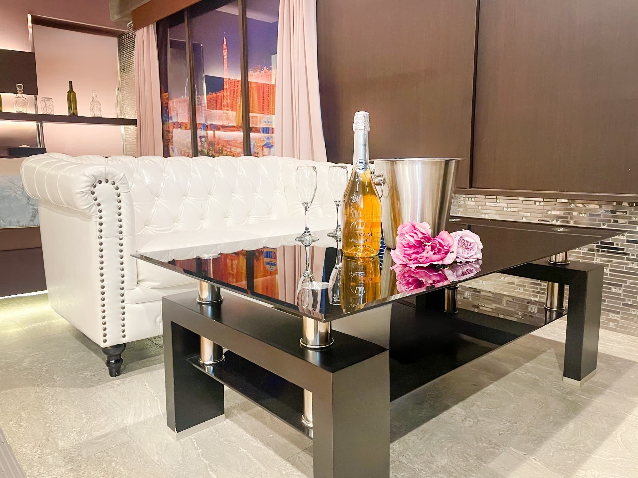 Una sala de estar moderna con un sofá de cuero blanco, una mesa de centro de vidrio con una botella y flores, y vistas a la ciudad a través de la ventana.