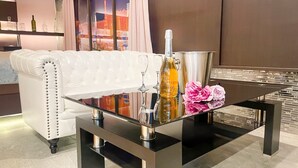 Una sala de estar moderna con un sofá de cuero blanco, una mesa de centro de vidrio con una botella y flores, y vistas a la ciudad a través de la ventana.