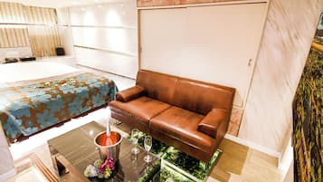 A modern hotel room with a bed, a brown leather sofa, a glass coffee table with a decorative item, and a small indoor garden.