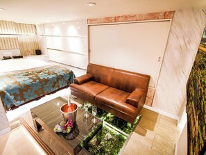 A modern hotel room with a bed, a brown leather sofa, a glass coffee table with a decorative item, and a small indoor garden.