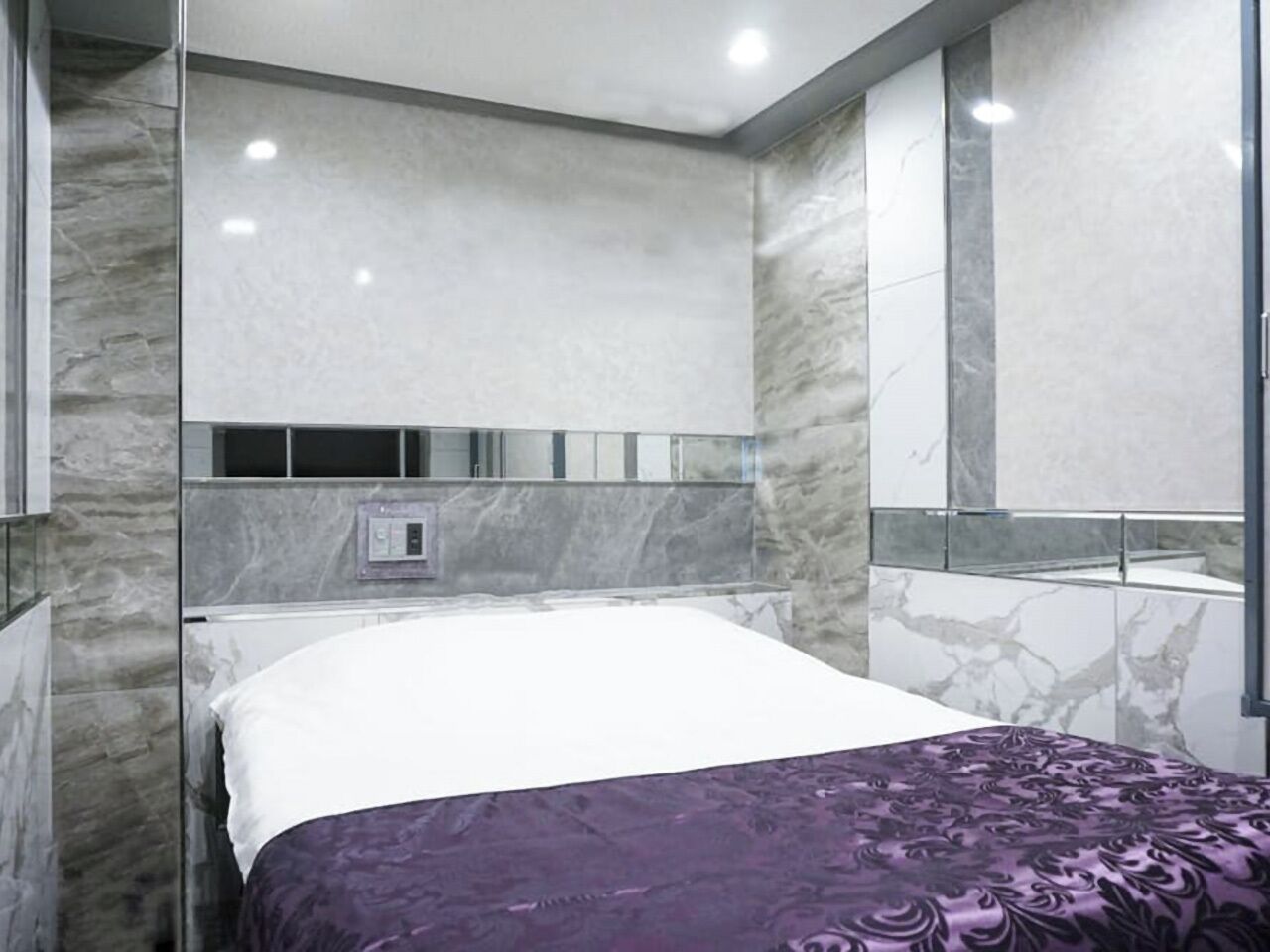 A modern hotel room with a marble wall, a bed with a white and purple bedspread, and a large mirror.