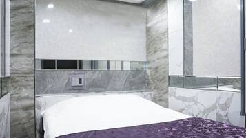 A modern hotel room with a marble wall, a bed with a white and purple bedspread, and a large mirror.