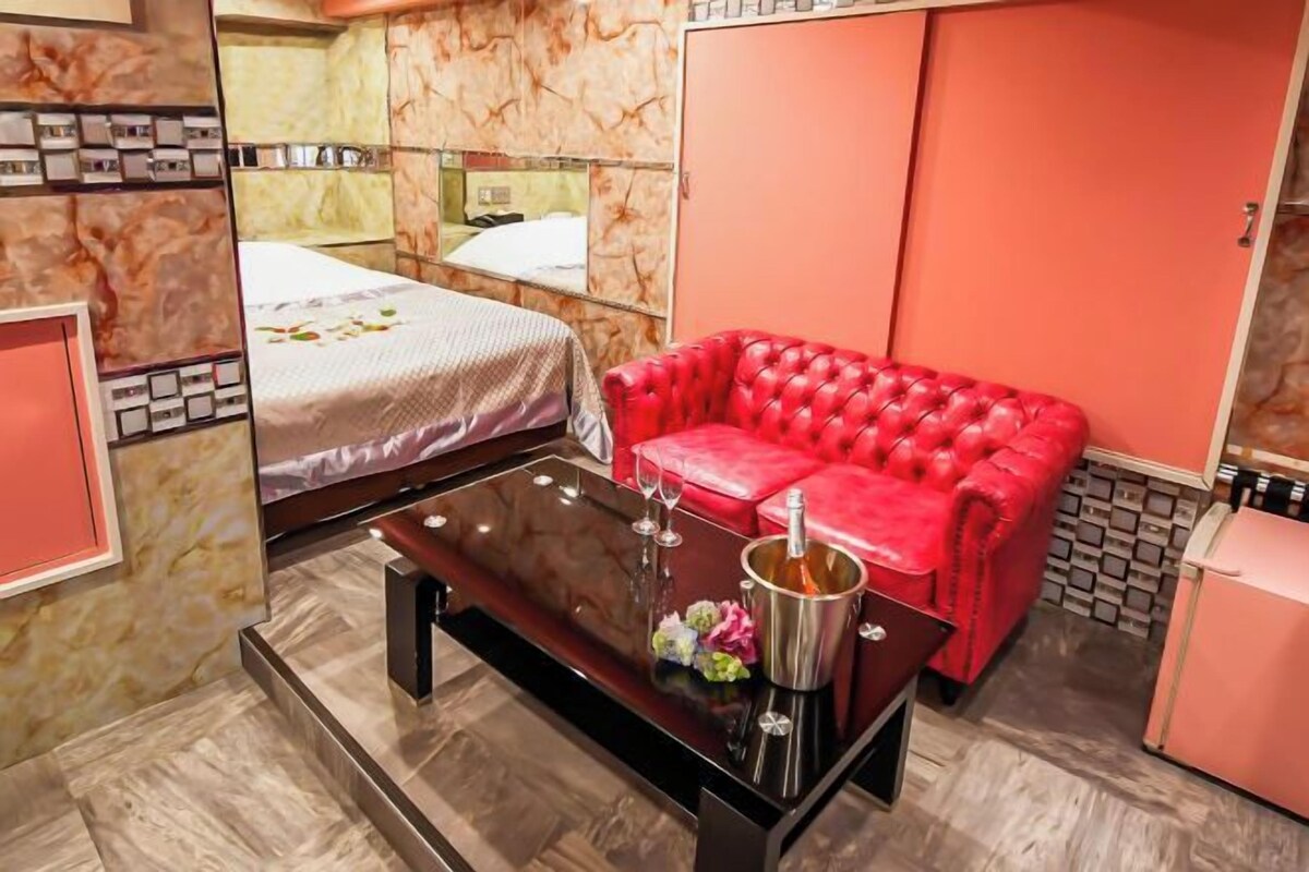 A hotel room with a bed, a red leather sofa, a small table with a bottle, and a pink refrigerator.