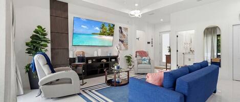 A modern living room with a large flat-screen TV, a blue sofa, a white recliner, and a coffee table.