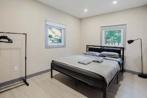 Ein Schlafzimmer mit einem Bett, zwei Fenstern, einer schwarzen Stehlampe und einem Kleiderständer.