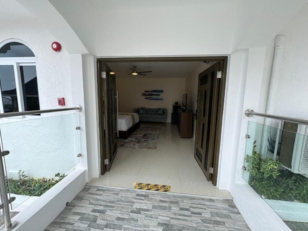 Entrada de hotel con barandilla de vidrio, detector de incendios, ventilador de techo, cama y un felpudo con la inscripción 'Precaución: Paso Reducción'.