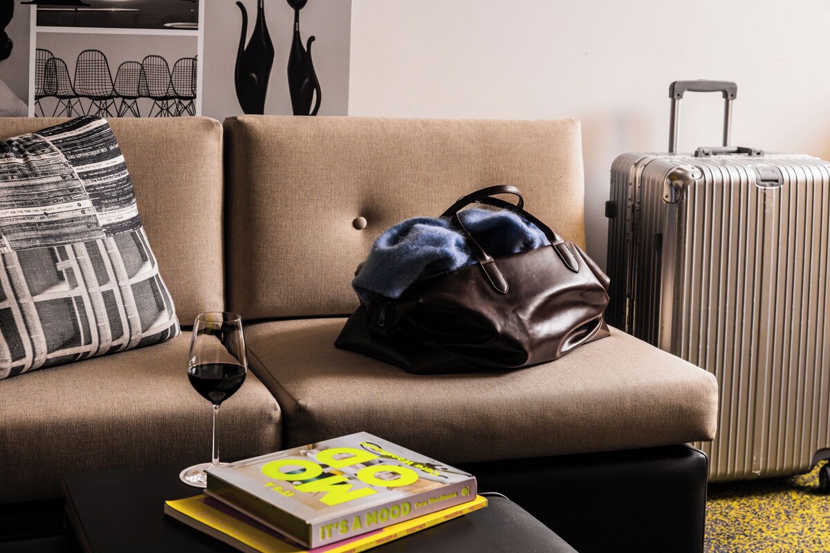 Un sofá color beige con un bolso de cuero marrón, un libro y una copa de vino tinto sobre una mesa negra.