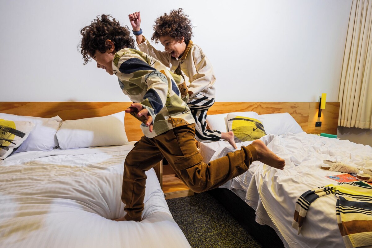 Dos personas saltando sobre una cama en una habitación con cabecera de madera, ropa de cama blanca y una mesita de noche visible.